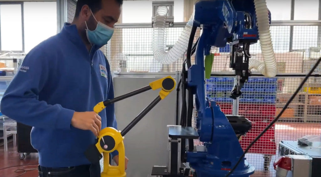 A man in an industrial setting wearing a blue shirt, standing next to a blue robotic arm, holding the stylus of a yellow MicroScribe 3D scanner.