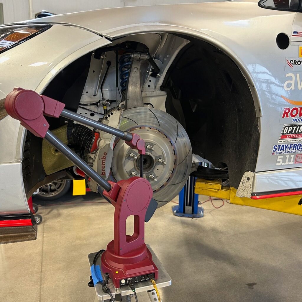 A red MicroScribe portable CMM on a stand measuring the front suspension of a white Nissan GT4 race car, with the wheel removed showing a slotted brake rotor, Brembo caliper, and blue coil-over spring.