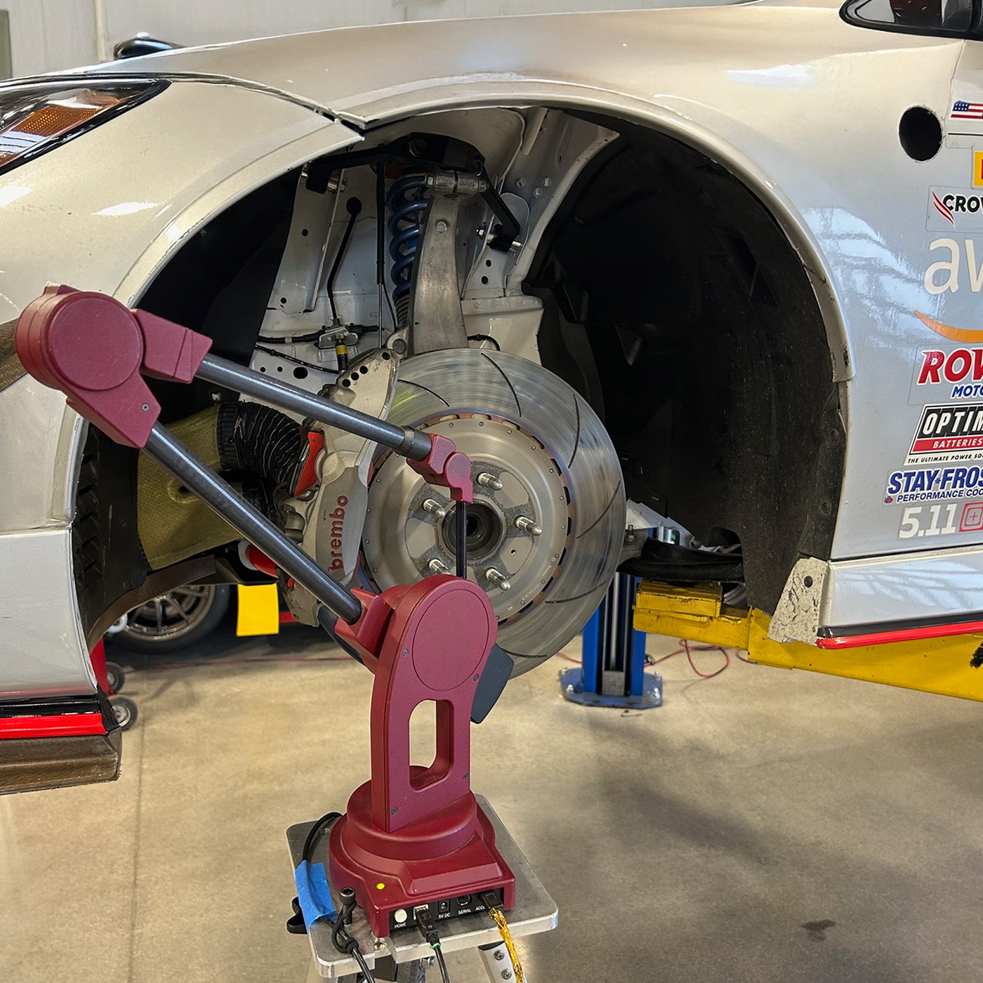 A red MicroScribe portable CMM on a stand measuring the front suspension of a white Nissan GT4 race car, with the wheel removed showing a slotted brake rotor, Brembo caliper, and blue coil-over spring.