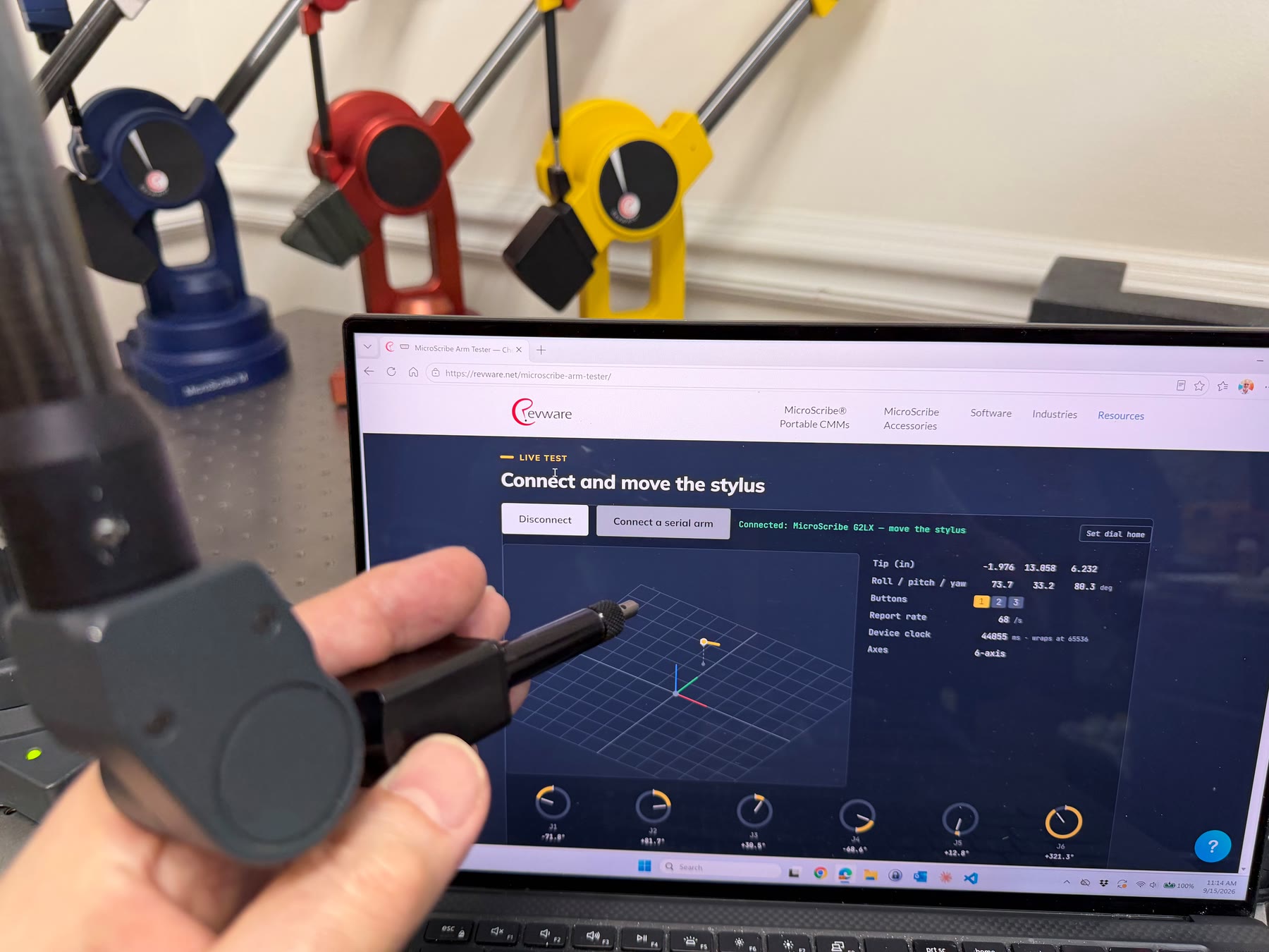 A hand holds the stylus of a MicroScribe arm in front of a laptop showing the MicroScribe Arm Tester page at revware.net. The page reads Connected: MicroScribe G2LX and shows a live stylus view, six joint dials, and position and orientation readouts. Blue, red, and yellow MicroScribe arms stand behind the laptop.