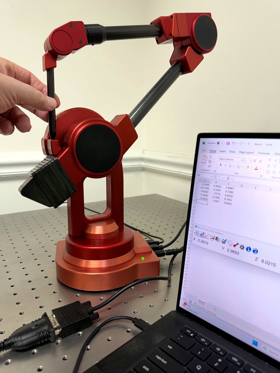 A hand moves the stylus of unit 0, the red 1995 MicroScribe prototype, while a Windows 11 laptop beside it shows live coordinates in MUS and captured points in Excel.